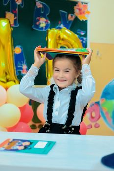 A cheerful schoolgirl holding books above her head in a decorated classroom with colorful balloons.