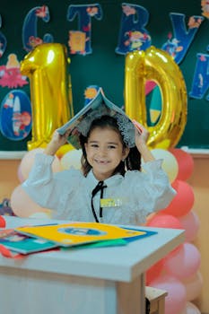 A happy schoolgirl in a classroom uses a book as a hat, celebrating her birthday with balloons and a cheerful atmosphere.