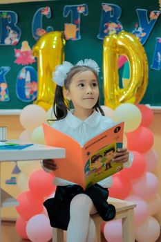Portrait of a schoolgirl sitting and holding a book in a colorful classroom setup with balloons.