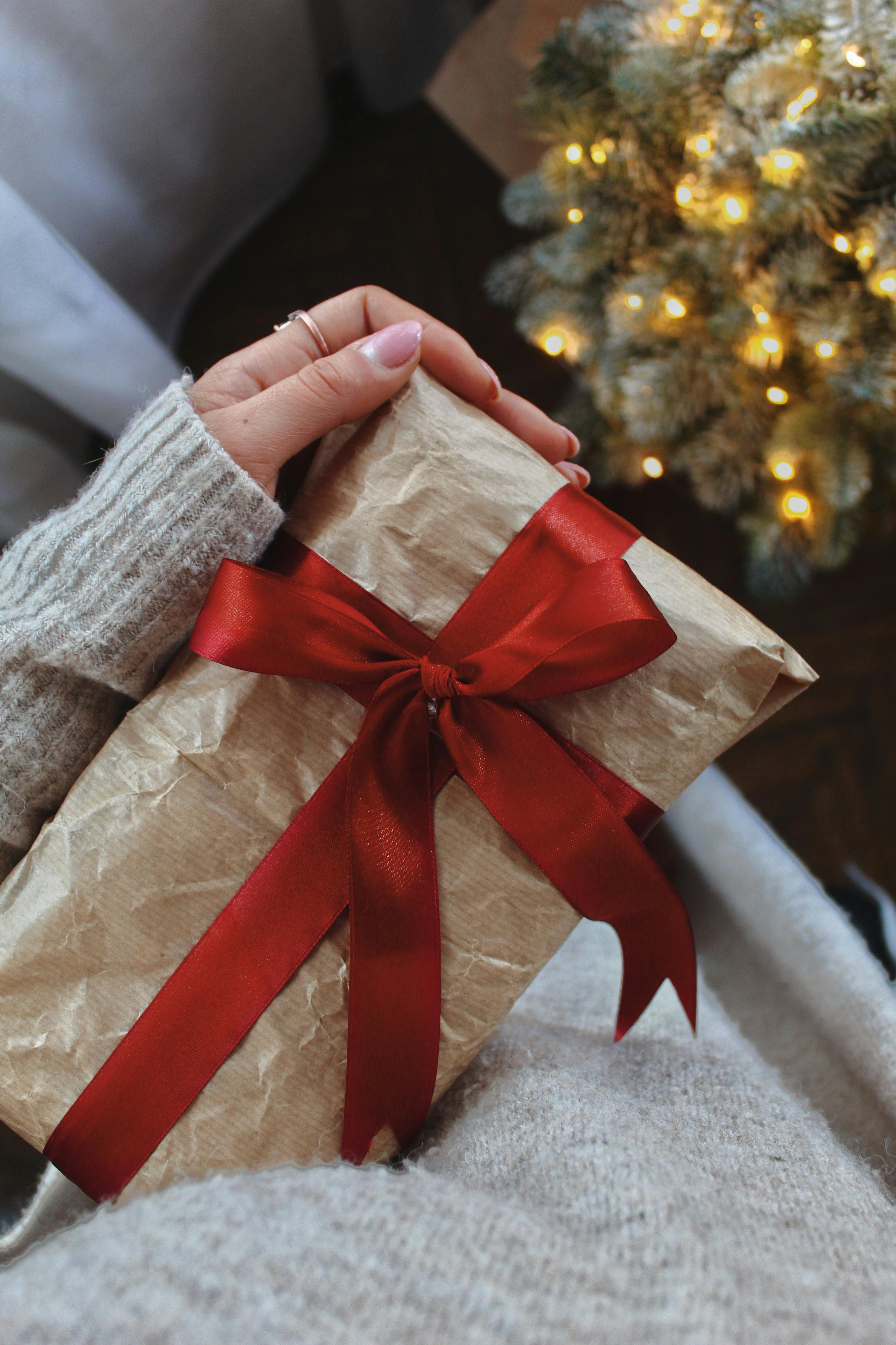 A cozy scene of a hand holding a Christmas gift wrapped with a red ribbon by a lit tree.