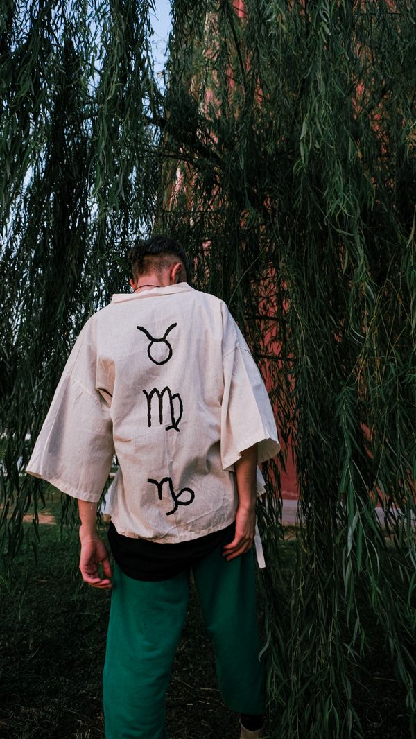 Free A man stands outdoors wearing a shirt with zodiac symbols, surrounded by willow trees. Stock Photo