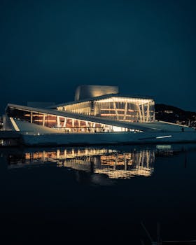 The striking Oslo Opera House glows warmly, reflecting in calm waterfront at night.