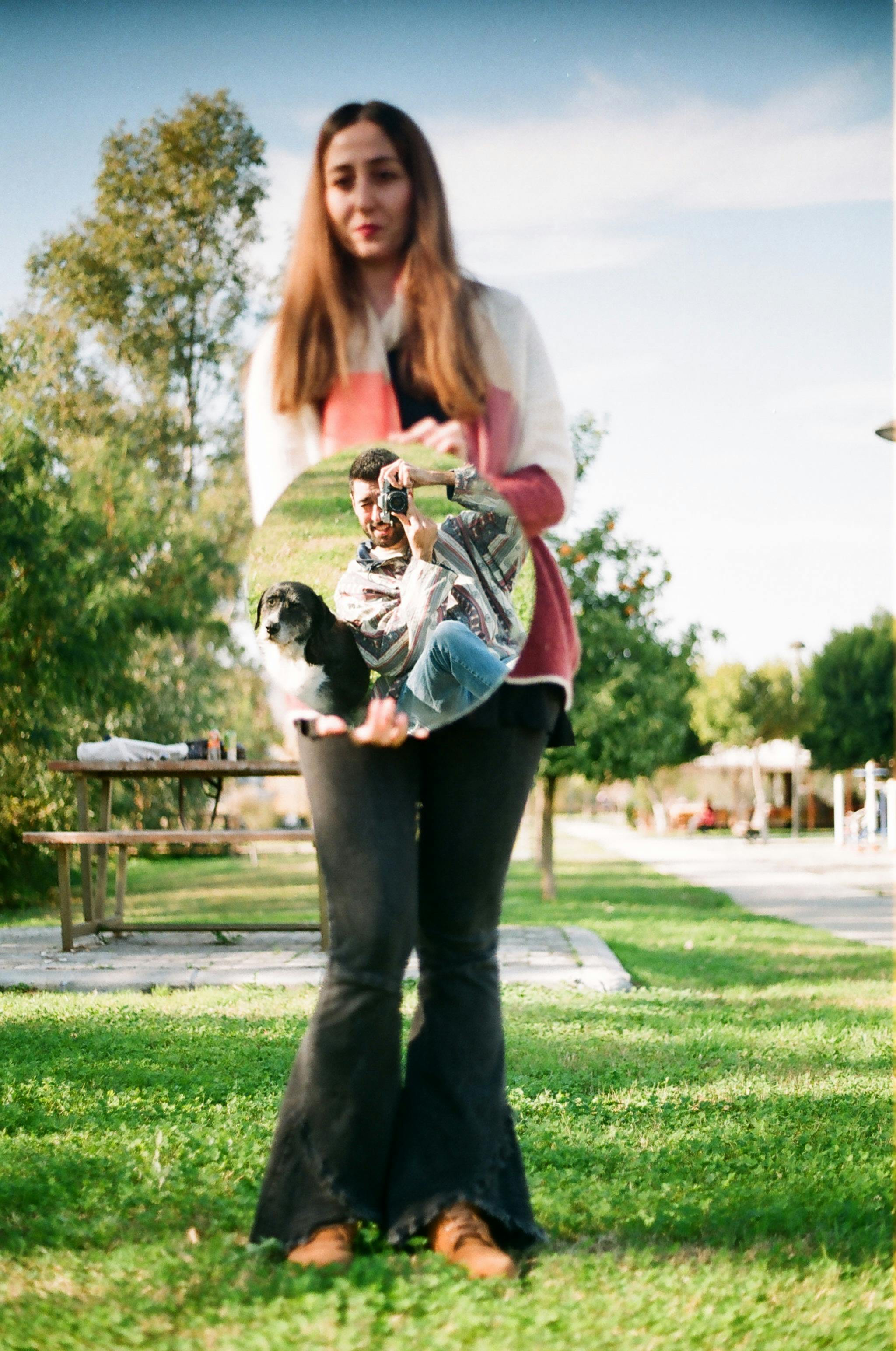 woman holding a mirror reflecting a man and a dog in a park