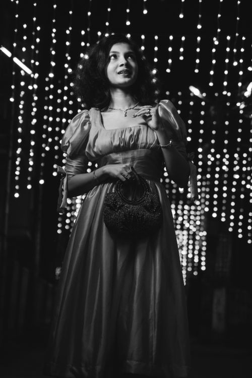 Free A stylish woman in an elegant gown poses under decorative string lights in this black and white fashion photo. Stock Photo
