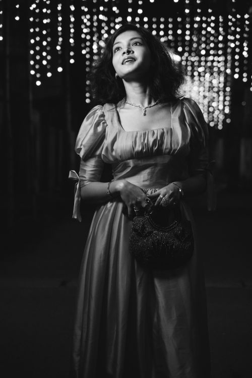 Free A young woman in a vintage satin dress posing confidently under twinkling string lights, holding a handbag. Stock Photo
