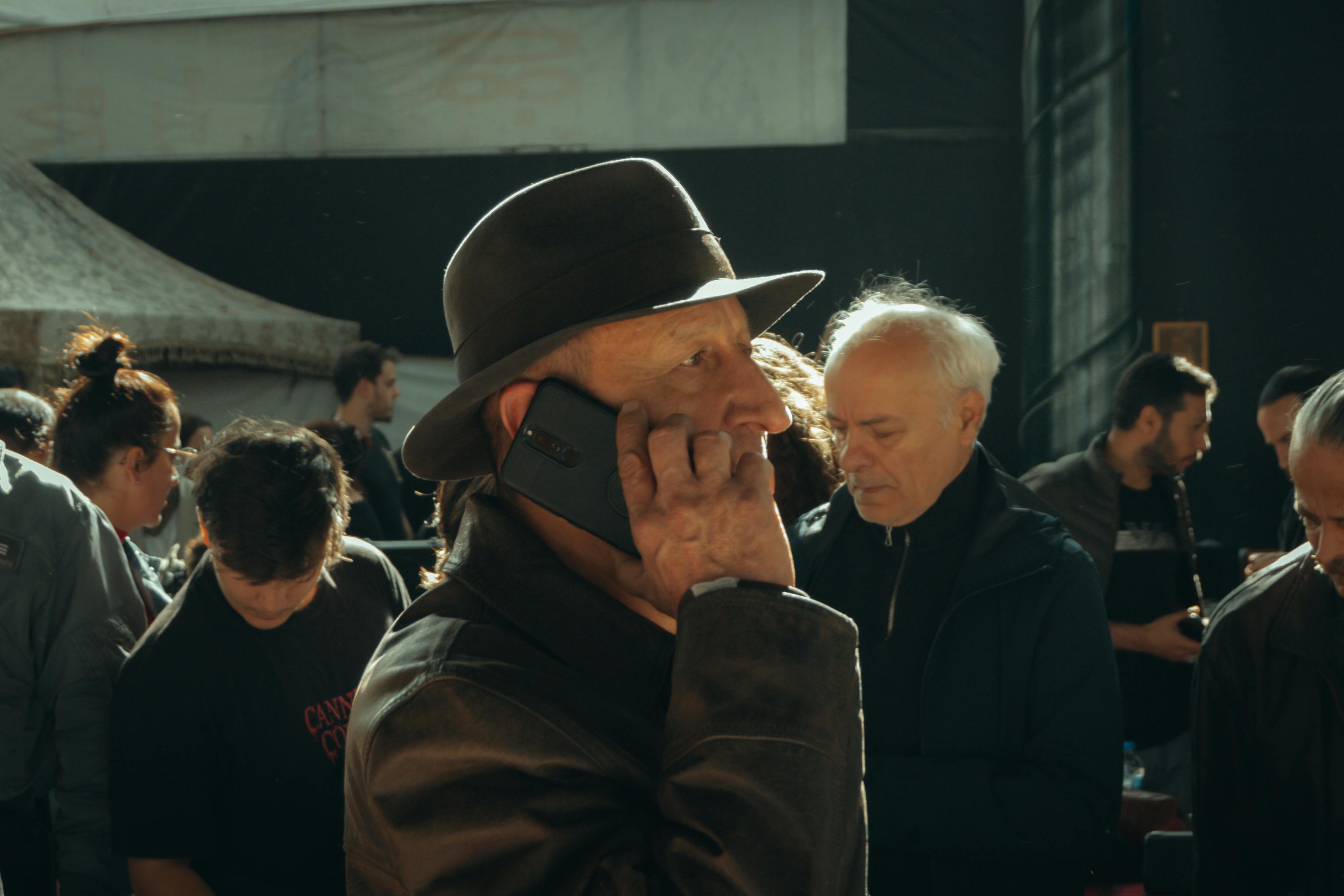 An elderly man in a hat talks on the phone amidst a busy street market setting.