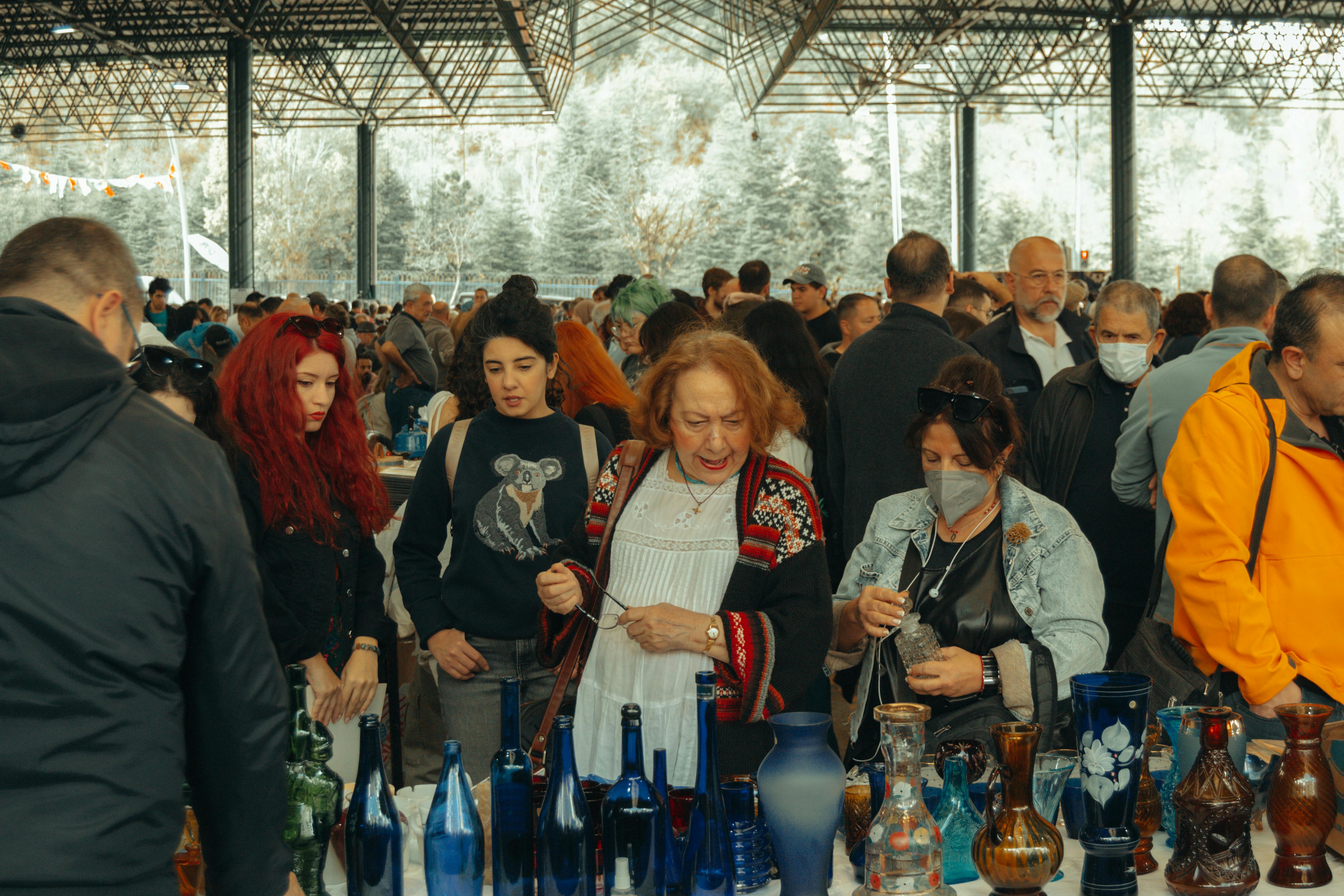 Crowd of people shopping for glassware at a bustling outdoor market under a pavilion.