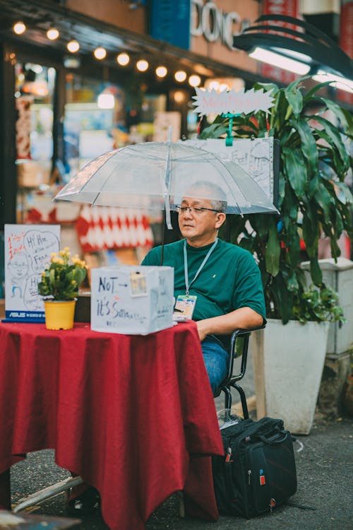 Man Sitting by the Table on the Sidewalk and Holding an Umbrella