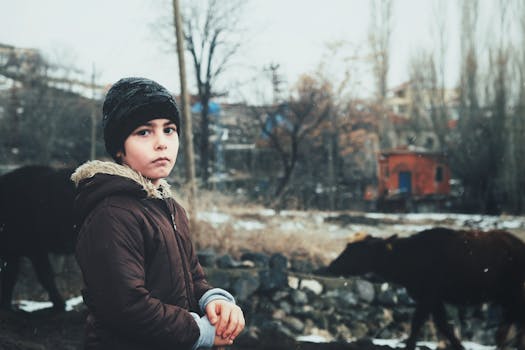 Young boy in winter clothing standing in rural village with cattle.