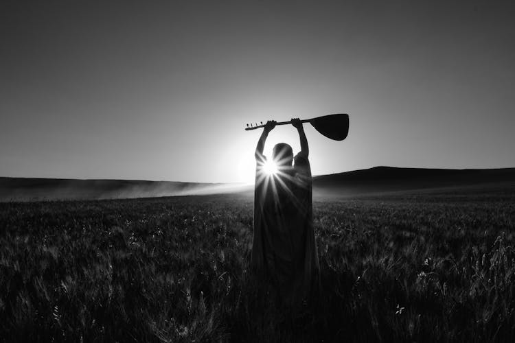 Black And White Photo Of Person Standing In Field With Balalaika Raised Above Their Head