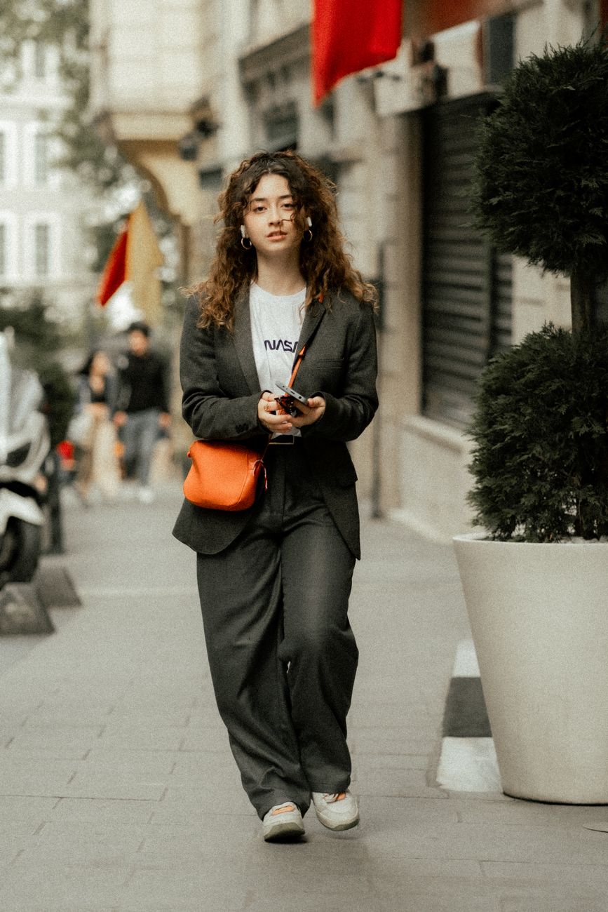 Casually dressed woman in fashionable attire walking along an urban sidewalk, carrying an orange purse.
