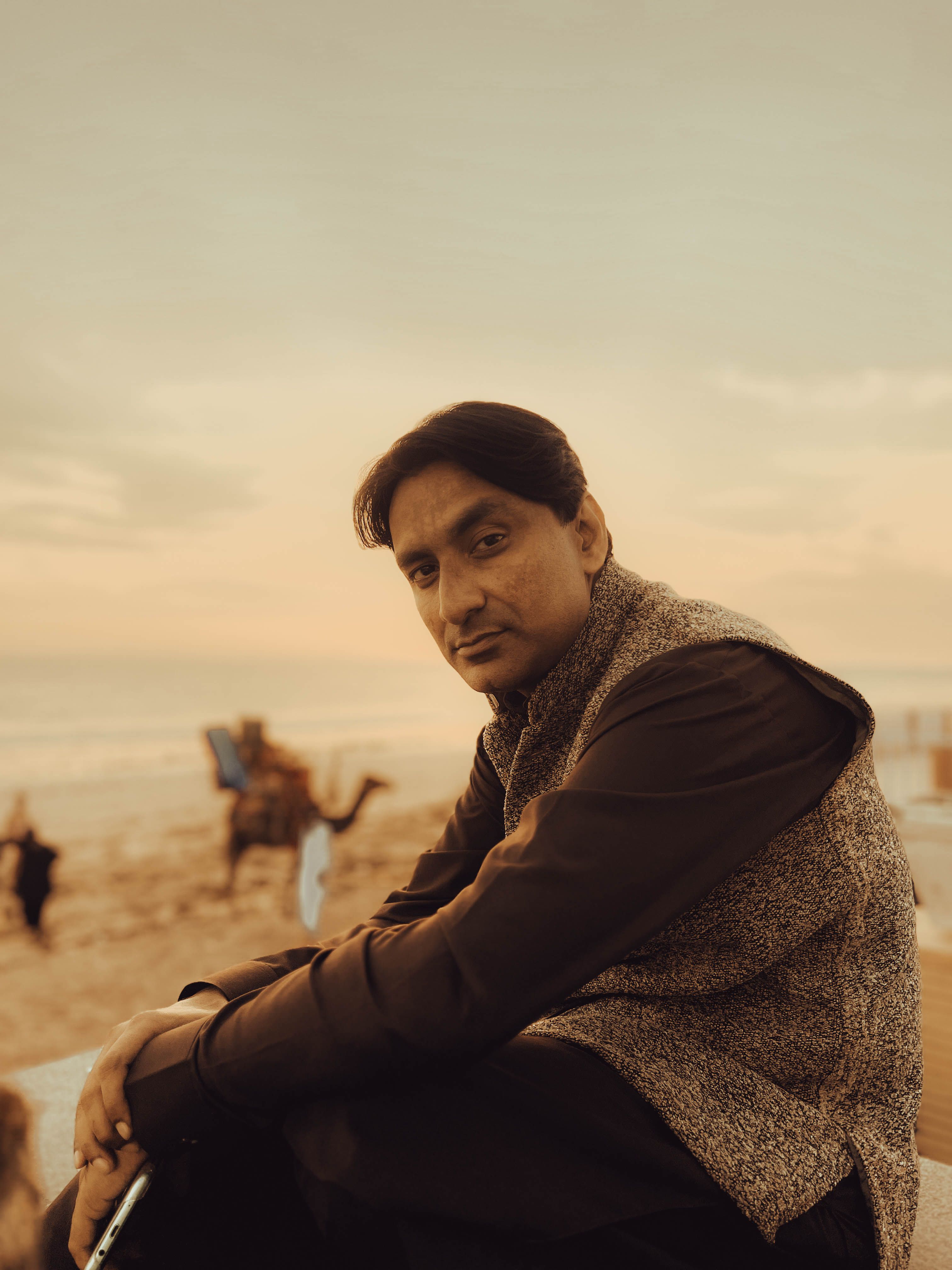 Man in a vest sitting by the beach at sunset, exuding calmness and style.