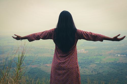 Free Back view of woman in pink dress standing on mountain, arms spread wide, embracing nature. Stock Photo
