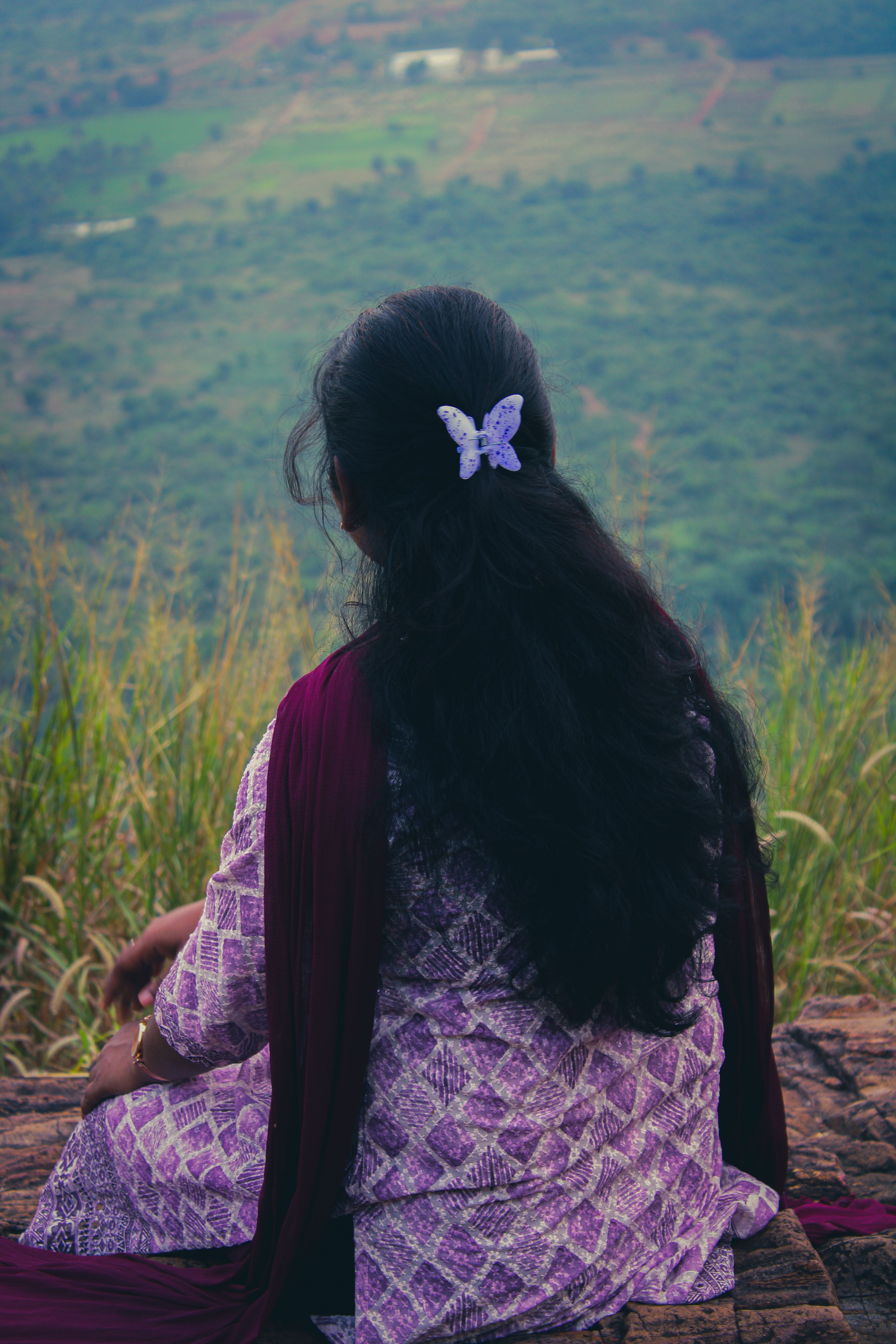 A woman with long black hair and a butterfly hairpin sits on a hill, enjoying a rural view.