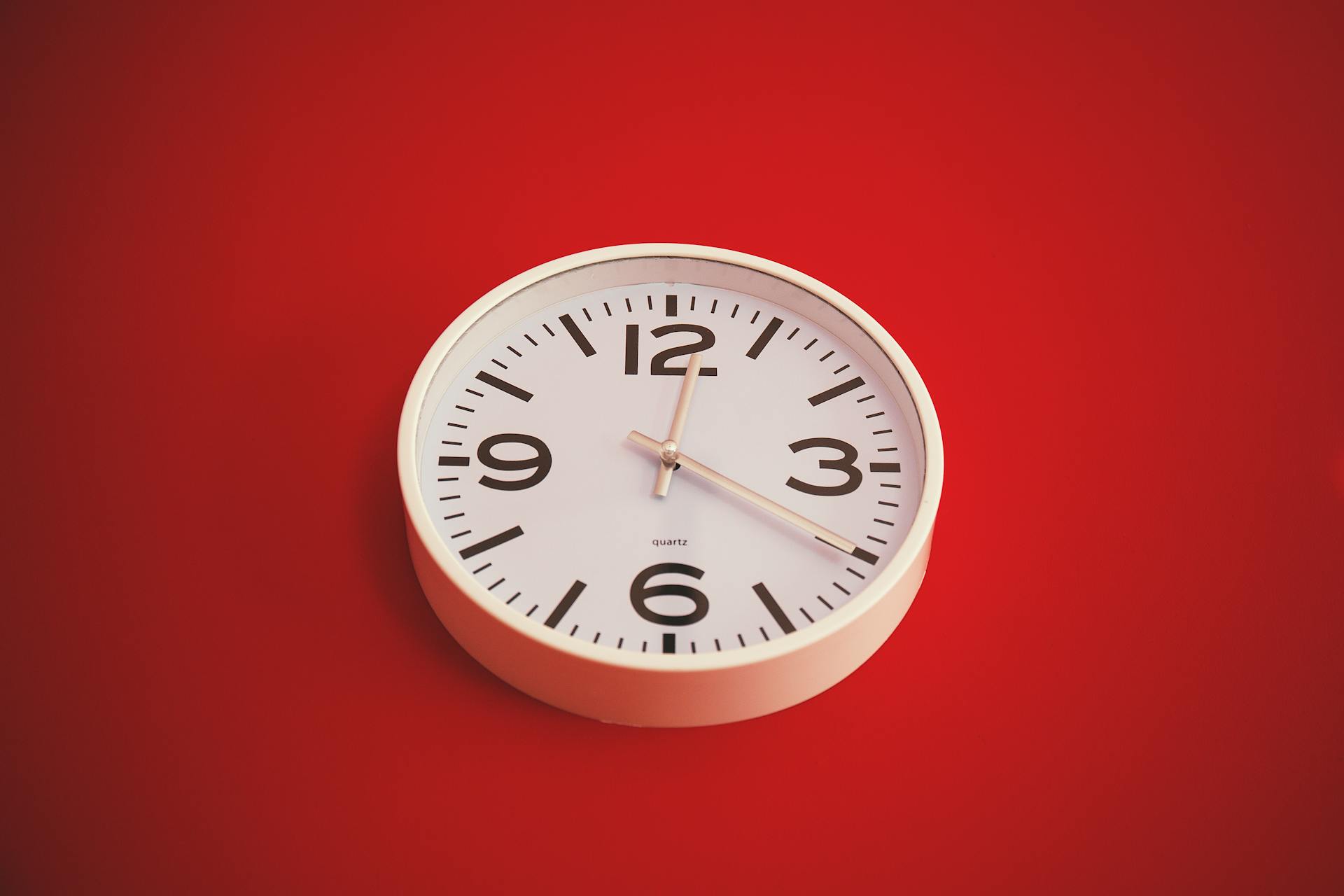 Minimalist round wall clock with a white face on a vibrant red background. Time marked as three o'clock.