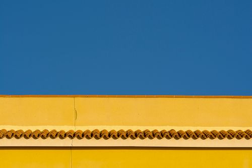 Free Minimalist photo of a yellow wall with a tiled roof contrasting against a clear blue sky. Stock Photo