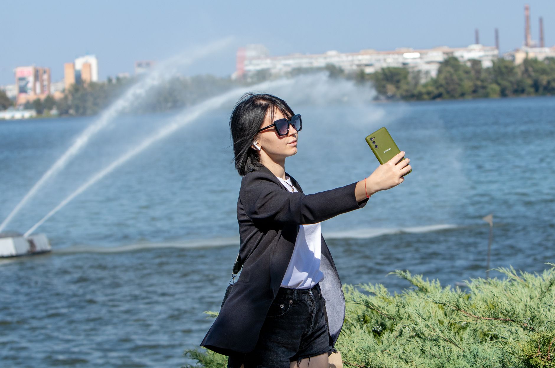 A woman in sunglasses takes a selfie by a fountain on a sunny summer day, capturing a vibrant outdoor scene.