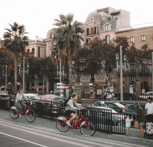 Free People cycling through the vibrant streets of Barcelona with palm trees and urban architecture in the background. Stock Photo