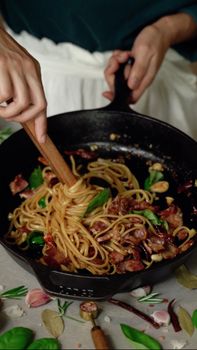 Close-up of hands preparing a savory pasta dish with bacon and herbs in a skillet.