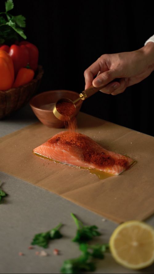 Free A hand sprinkles paprika on a fresh salmon fillet on baking paper for a vibrant culinary scene. Stock Photo