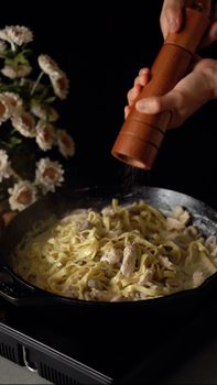 Close-up of hand seasoning creamy chicken pasta with pepper in a skillet.