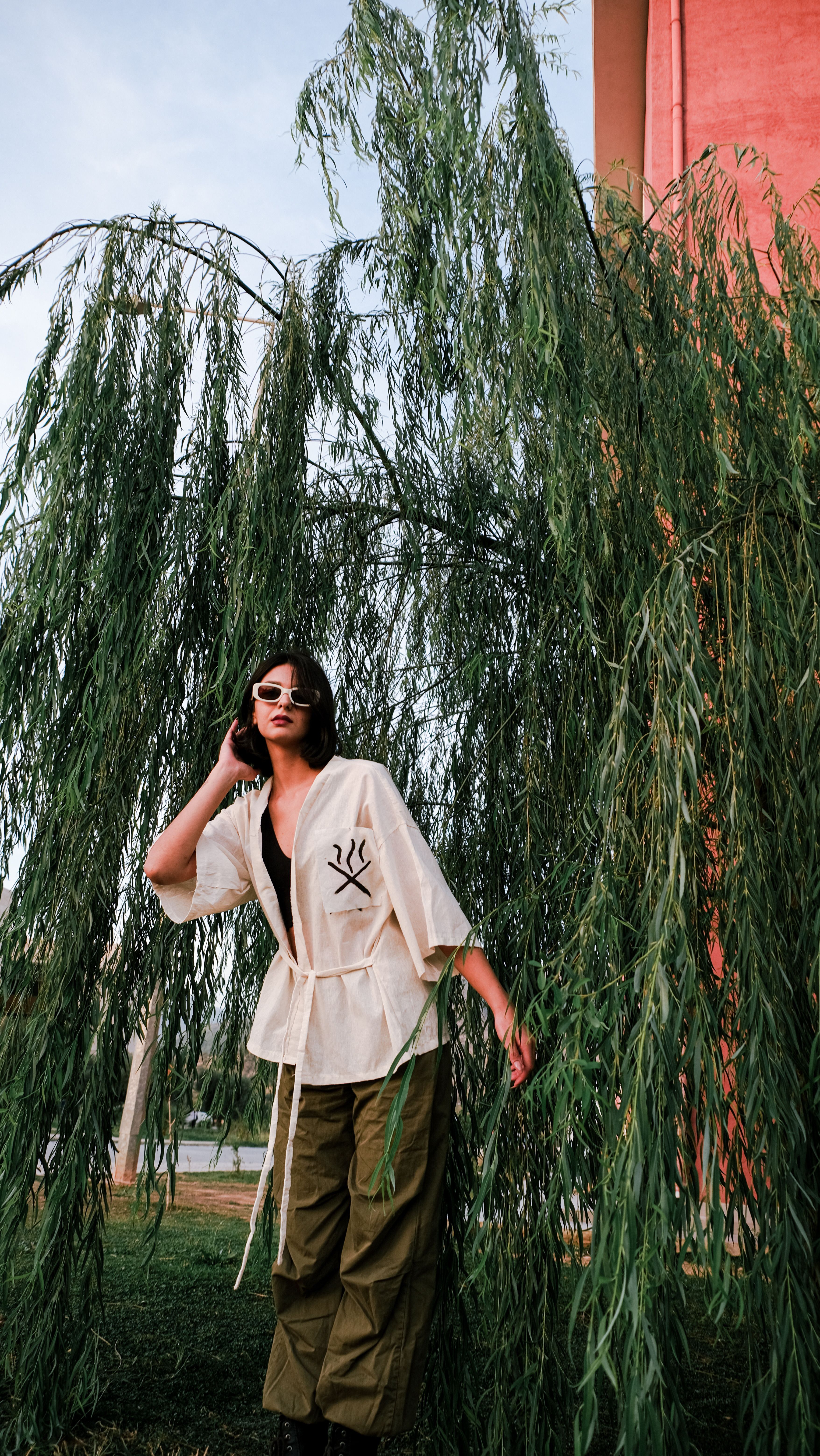 Chic woman in sunglasses poses by a weeping willow tree, showcasing modern style and nature's beauty.
