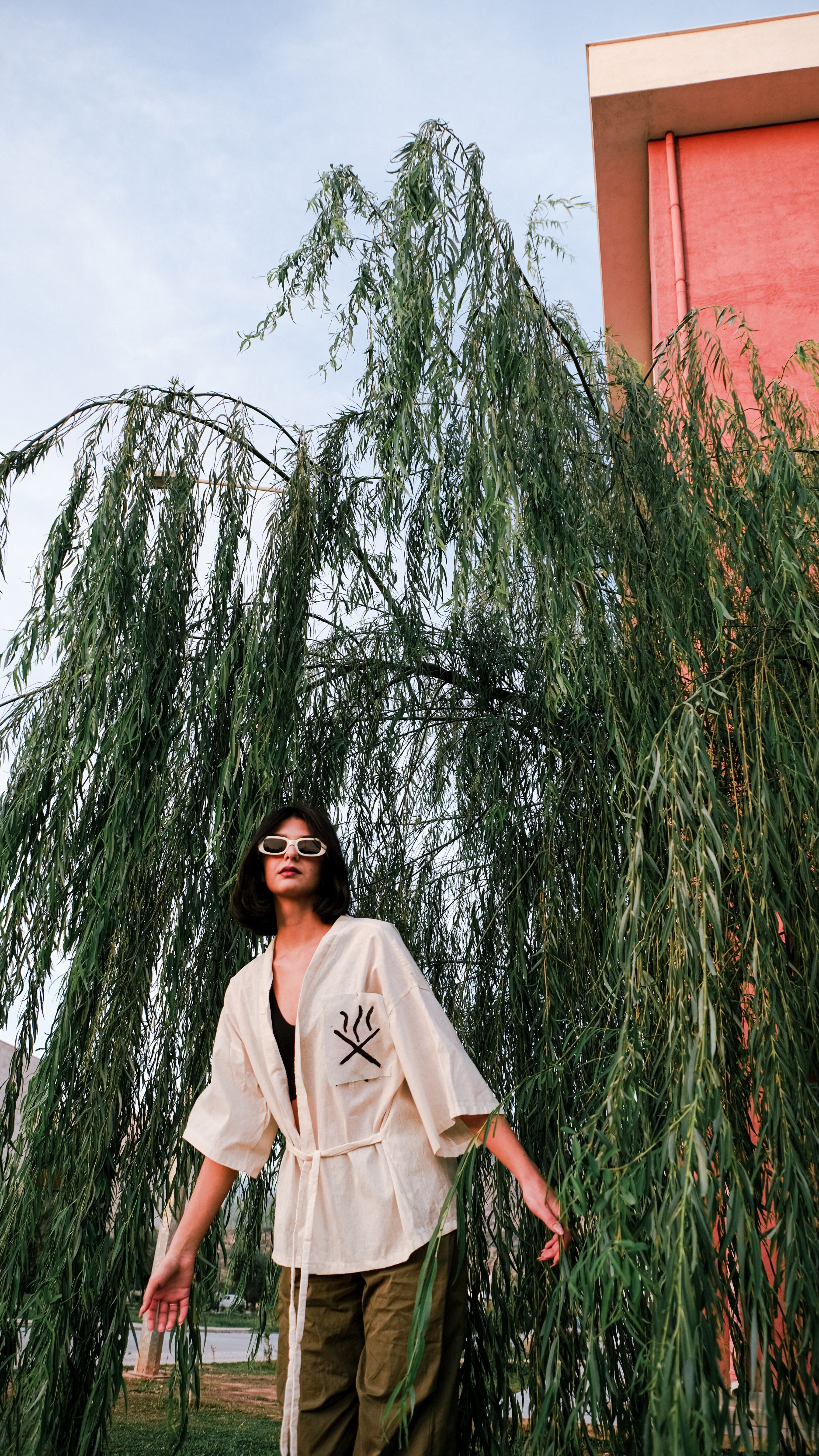 Stylish woman with sunglasses poses near lush greenery and a building, showcasing modern fashion.