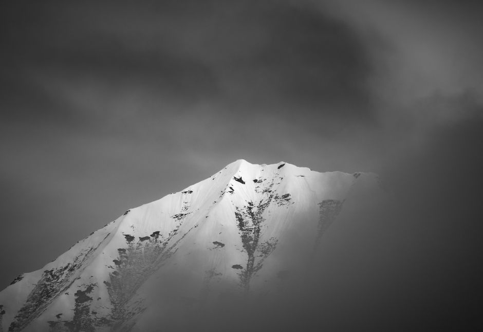 Black and white photo of a snow-covered mountain peak enveloped in mist, creating a dramatic and moody atmosphere.