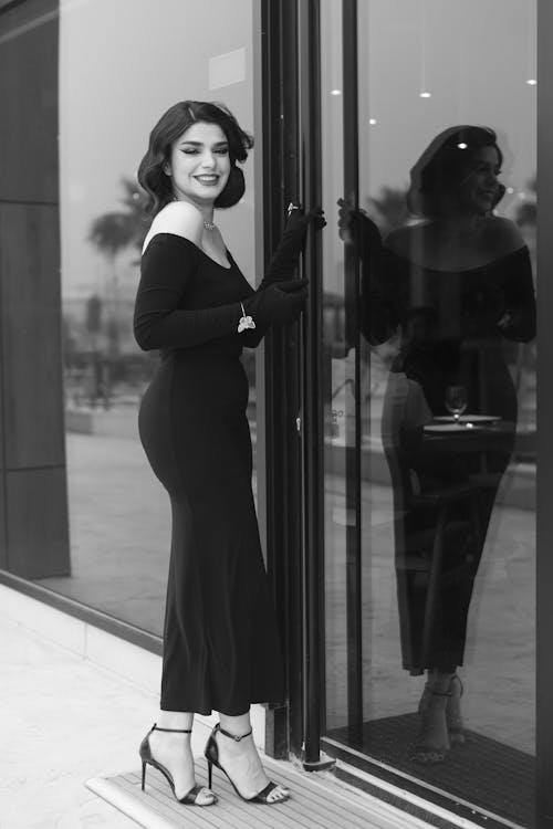 Young Elegant Woman in a Long Black Dress and High Heels Standing by the Window and Smiling 