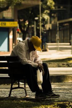 An elderly man with a yellow cap sitting alone on a park bench, looking thoughtful.