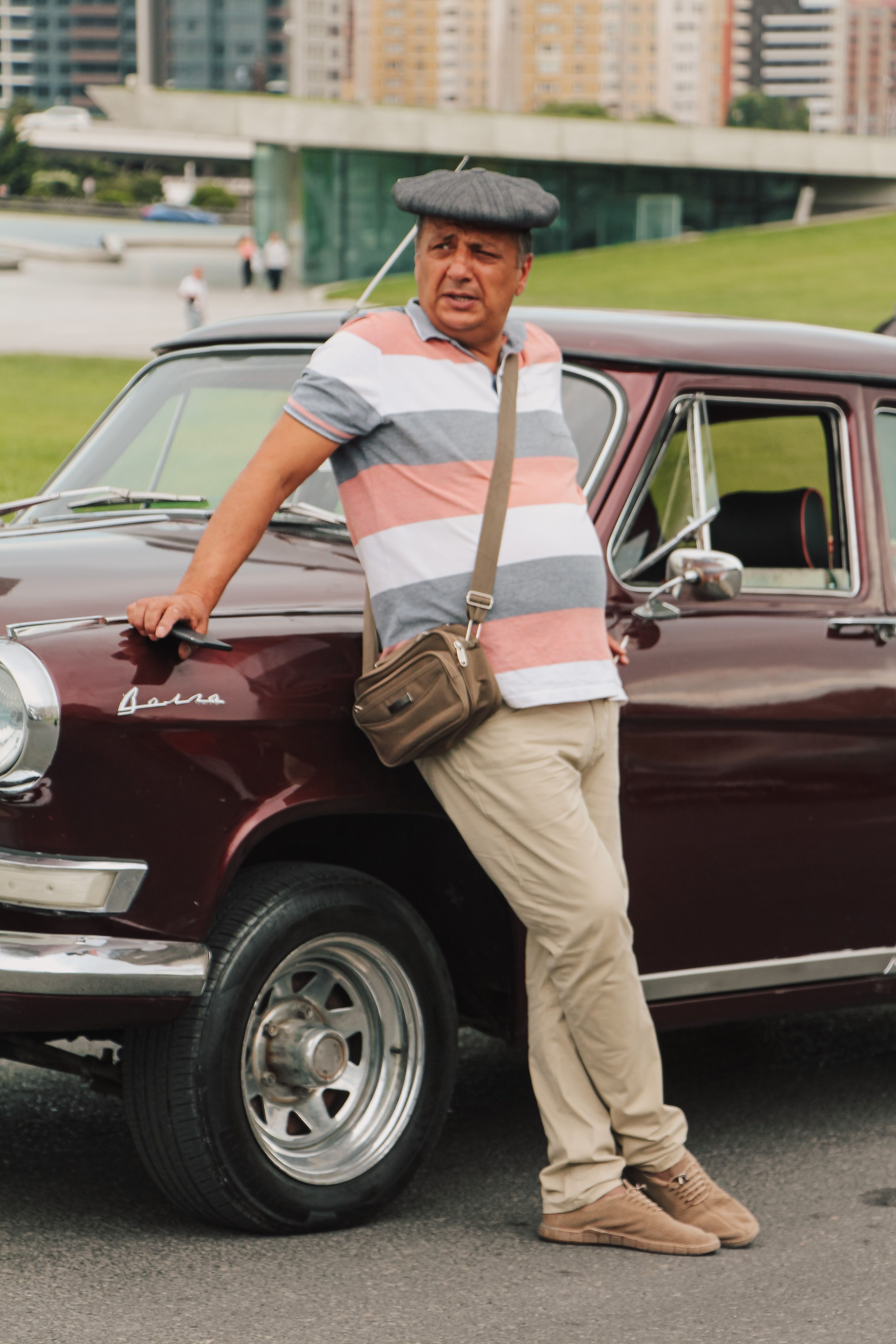 A man in a hat leans against a vintage car in an urban setting in Baku, Azerbaijan.