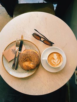 A warm and inviting breakfast table with cappuccino, bagel, and sunglasses.