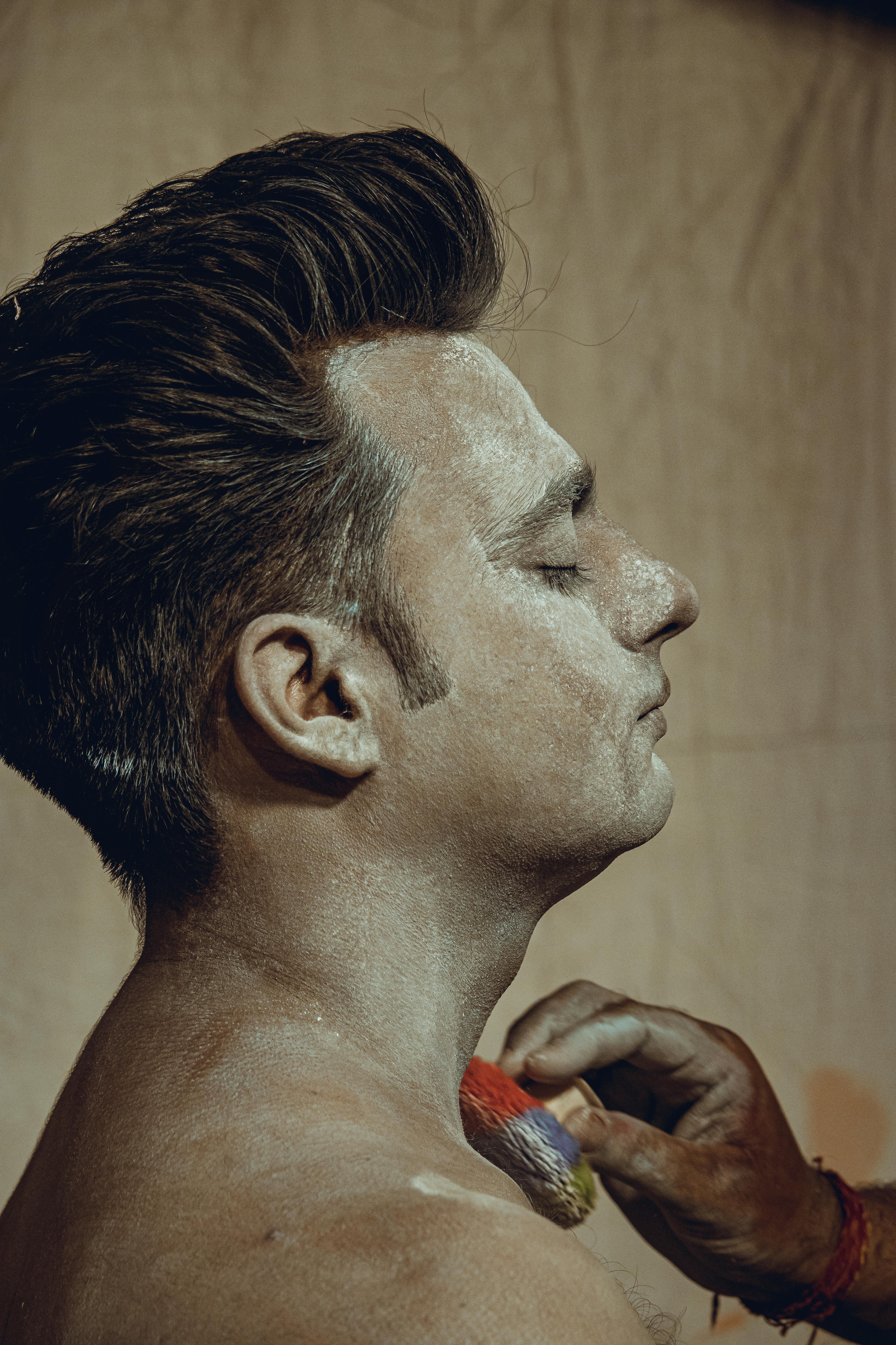 A man applying makeup with a brush, eyes closed, in a studio setting.