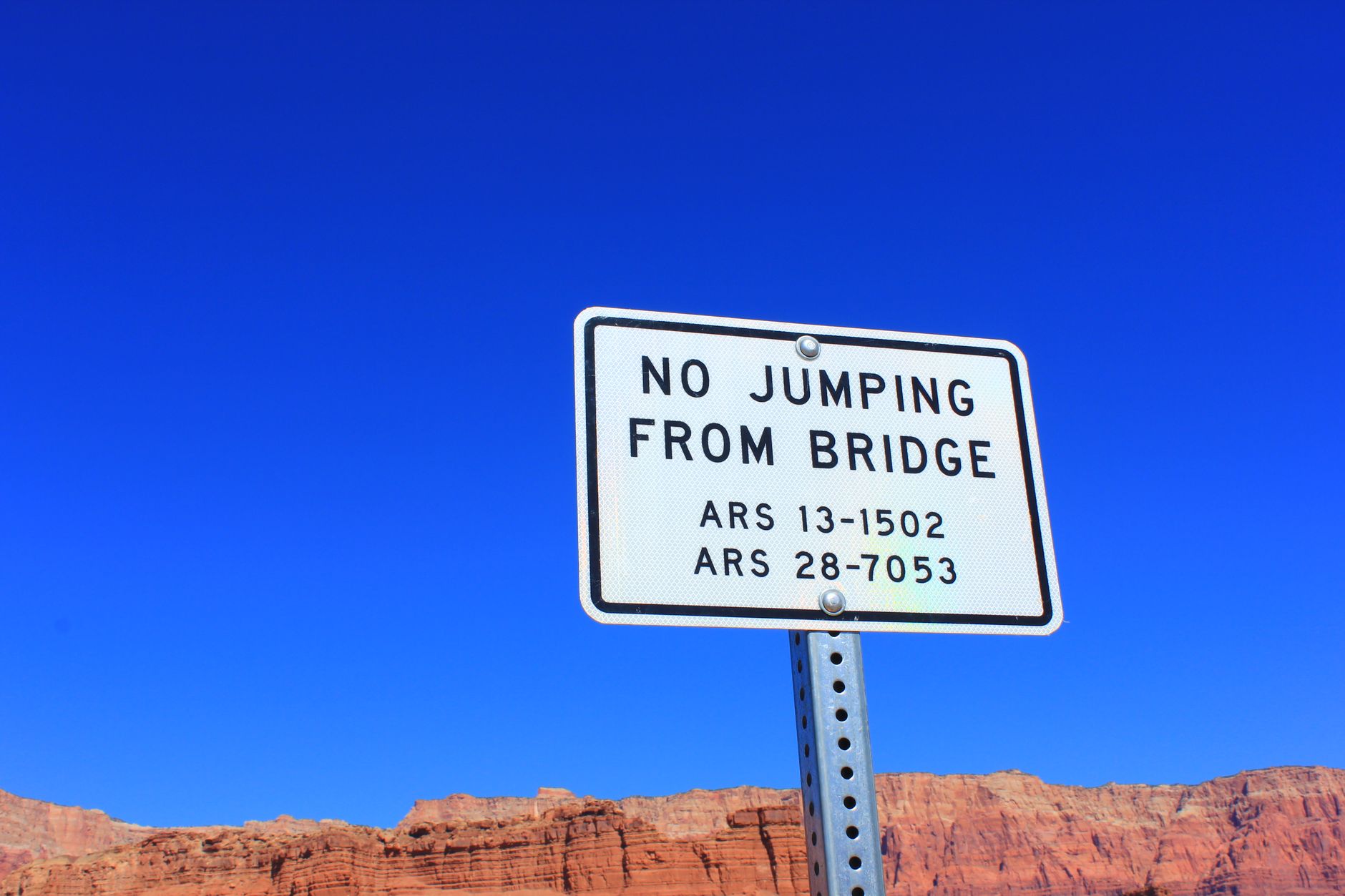 A warning sign stating 'No Jumping from Bridge' against a clear blue sky and rugged terrain.