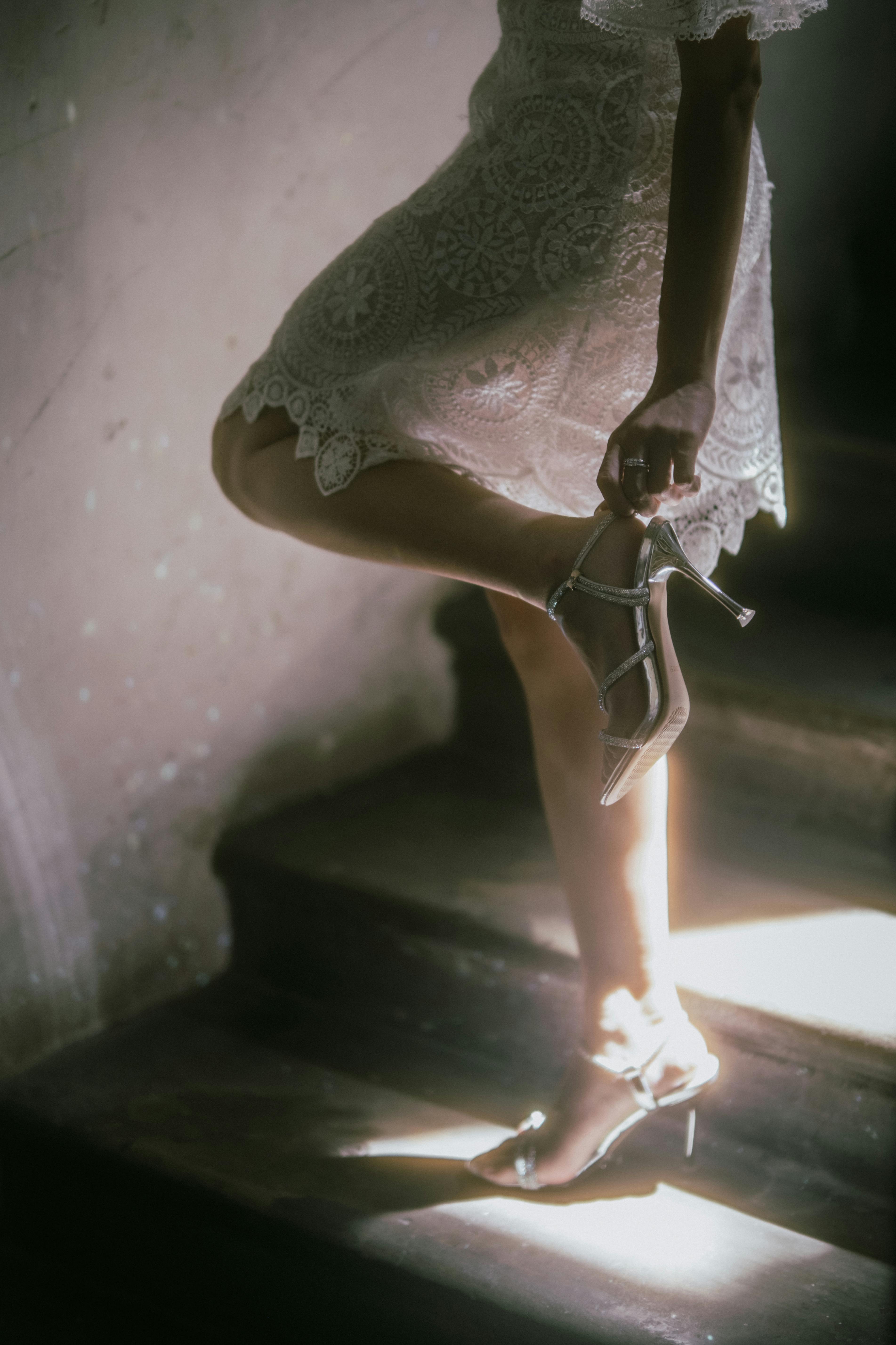 A bride in a lace dress gracefully descends dimly lit stairs, holding silver heels.