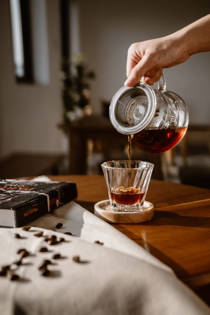 Hand pouring hot coffee into a glass using a glass kettle on a wooden table with scattered coffee beans.