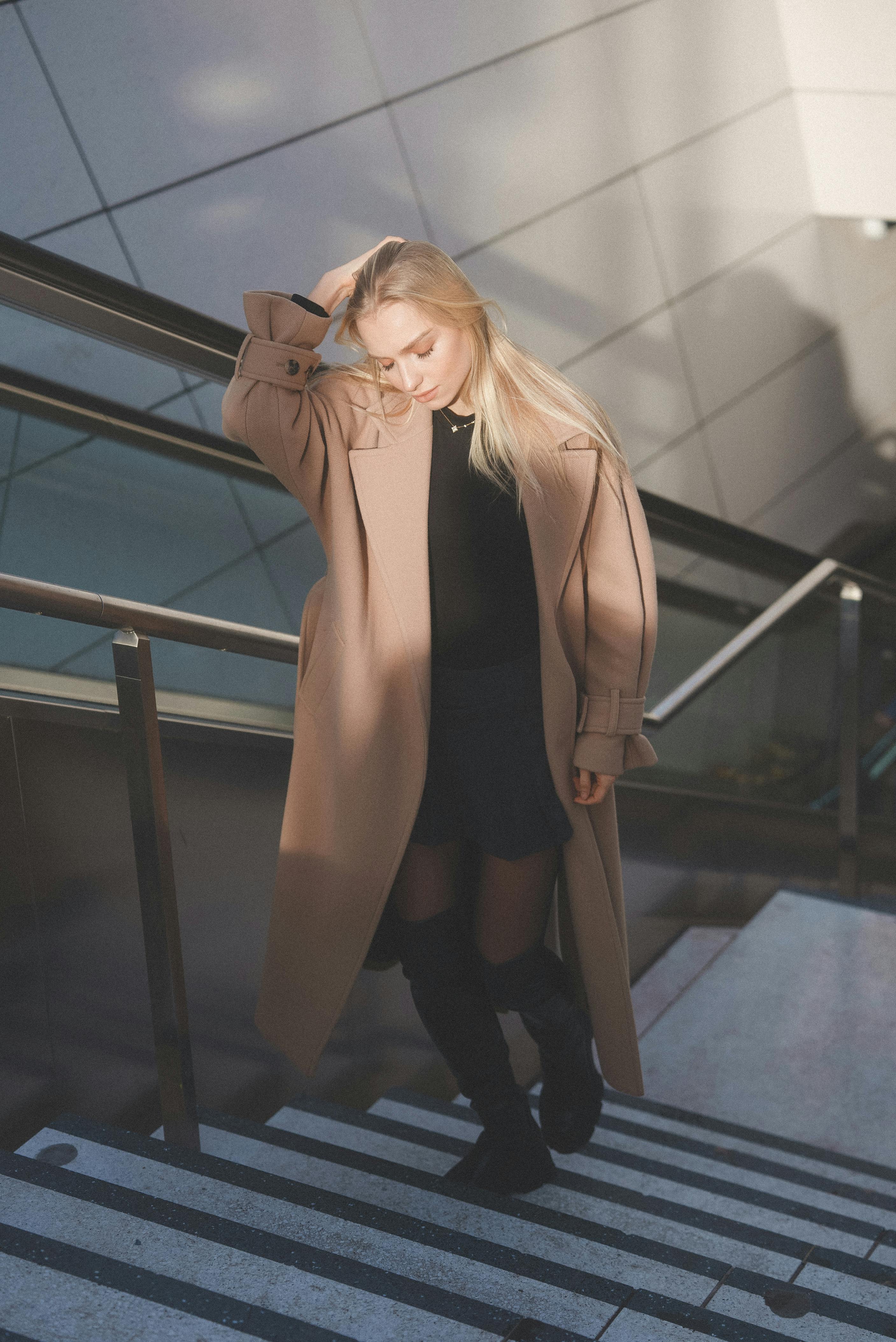Elegant woman in stylish beige coat walking down modern architectural stairs in daylight.