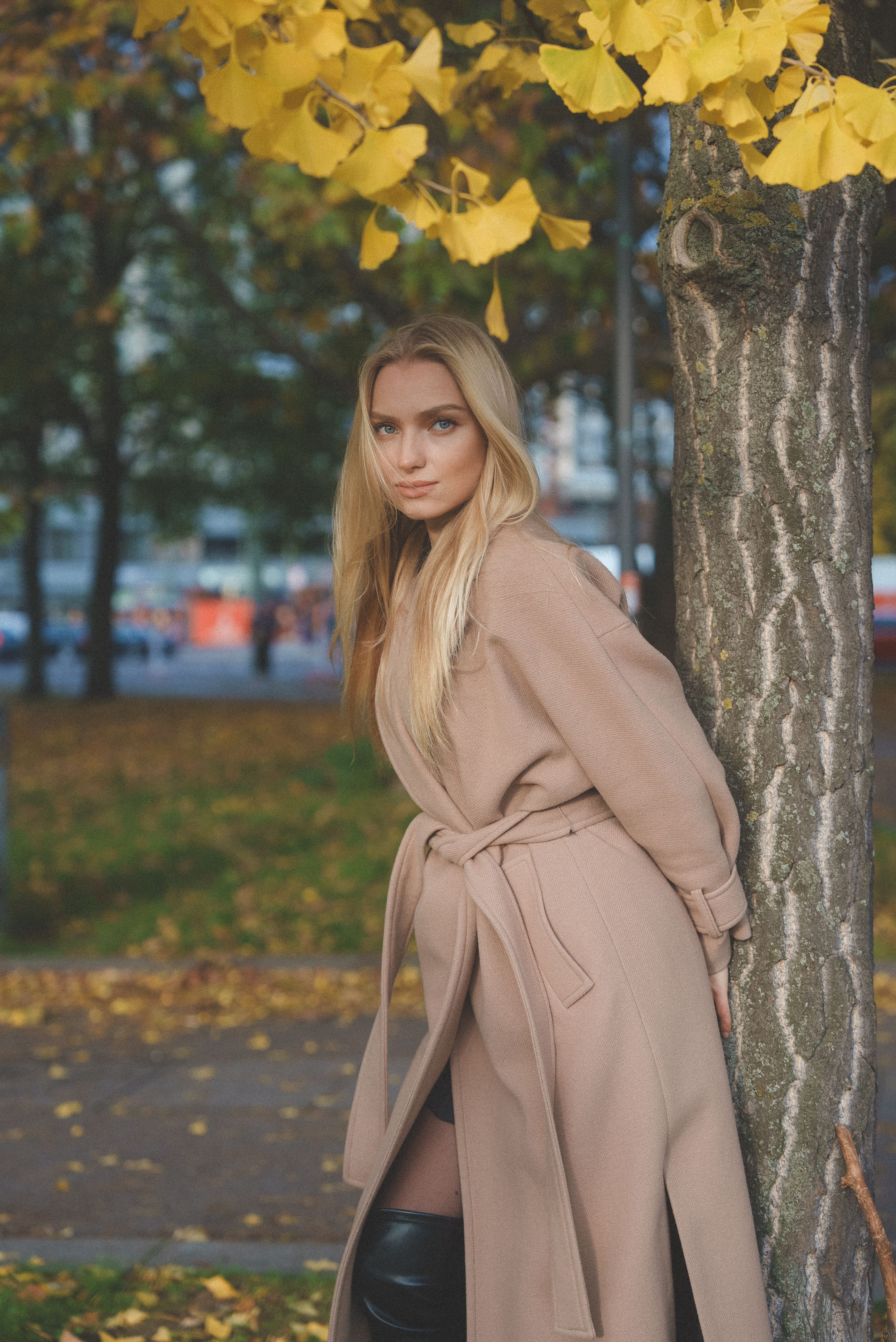 Portrait of a blonde woman in a stylish coat leaning against a tree in a vibrant autumn park.