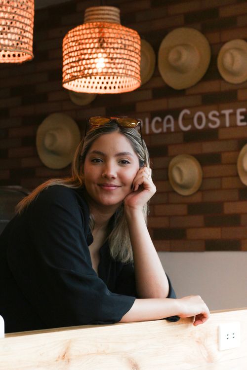 Free Young woman leaning on counter, smiling in a cozy hat-themed café with warm lighting. Stock Photo