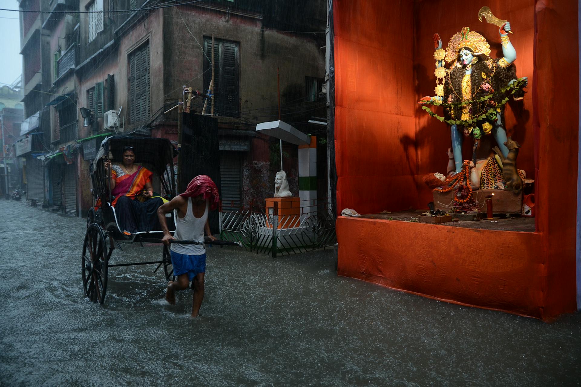 A rickshaw puller transports a passenger through flooding in Kolkata near a Kali statue during heavy rain.