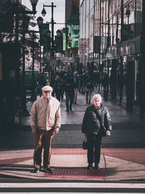 Free Elderly couple standing at a crosswalk in a bustling city street with pedestrians. Stock Photo