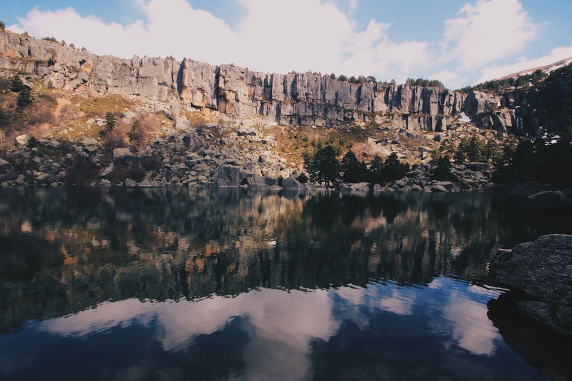 бесплатная Серая скалистая гора возле водоема под голубым небом с белыми облаками в дневное время Стоковое фото