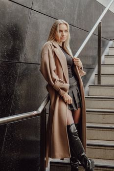 Stylish woman in a trench coat and leather boots poses on an urban staircase.