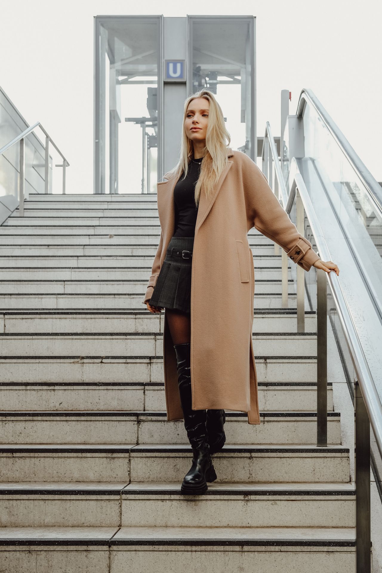 Long camel coat worn over a short dress with knee-high boots on city stairs