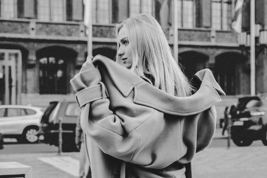 Elegant blonde woman posing on city street, enveloped in a coat with urban backdrop in black and white.