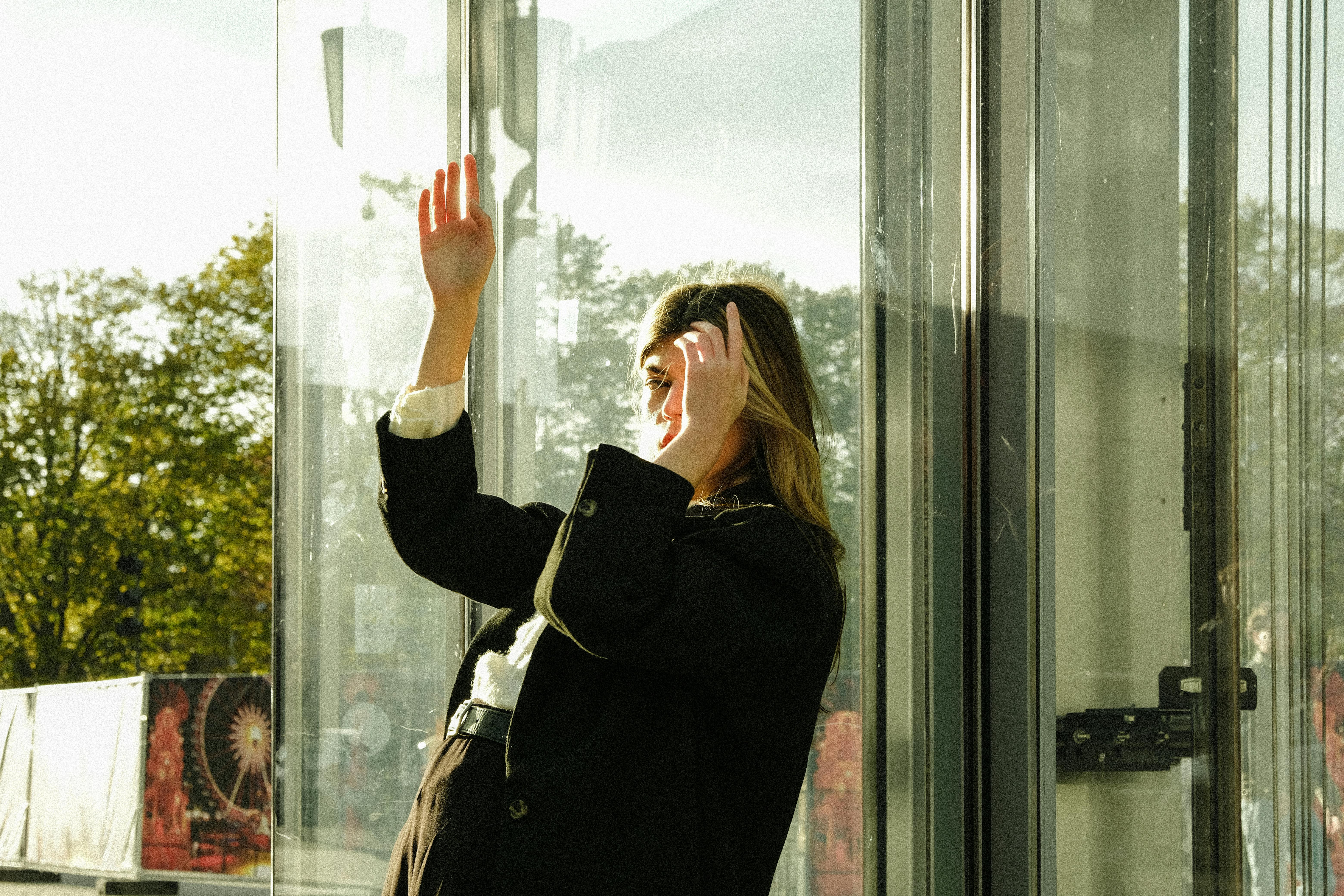 Free Fashionable woman in black coat poses elegantly by a glass window with sunlight. Stock Photo