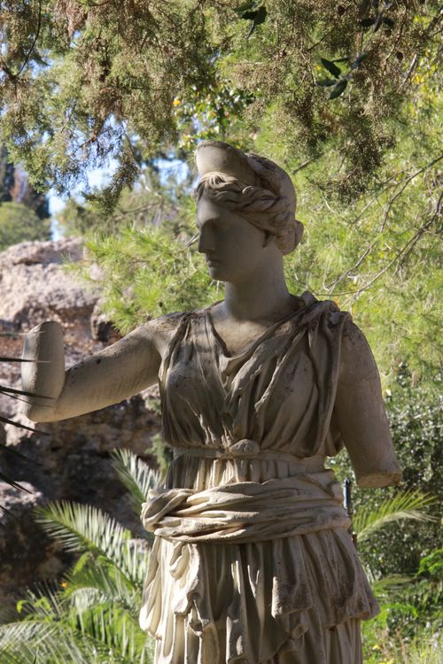 Free Classical stone sculpture of a Greek goddess in an outdoor park, surrounded by greenery. Stock Photo