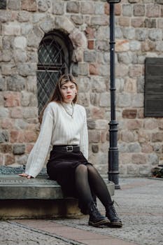 Stylish woman posing outdoors in urban setting with white sweater and black skirt.
