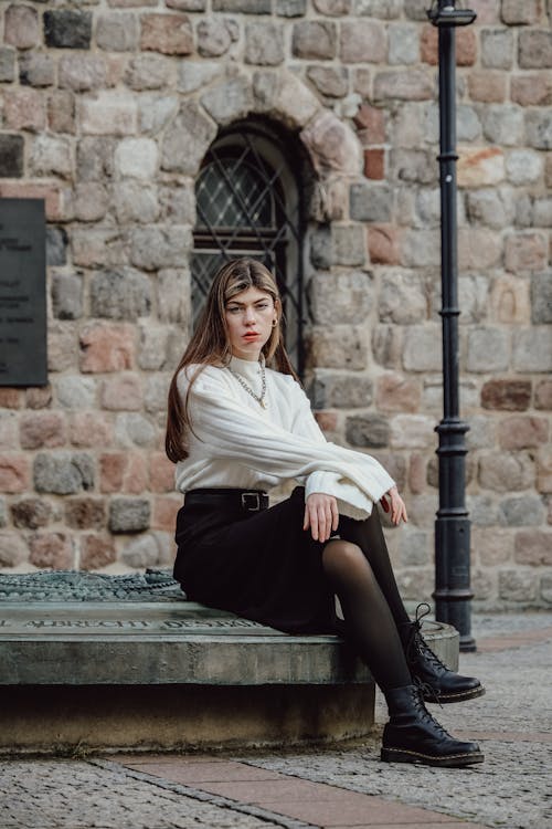 Model in a White Wool Cuffless Sweater and a Black Skirt Sitting in the City Square