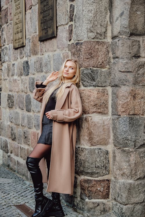 Model in a Beige Coat over a Black Blouse and a Gray Pleated Mini Skirt Leaning Against a Stone Wall